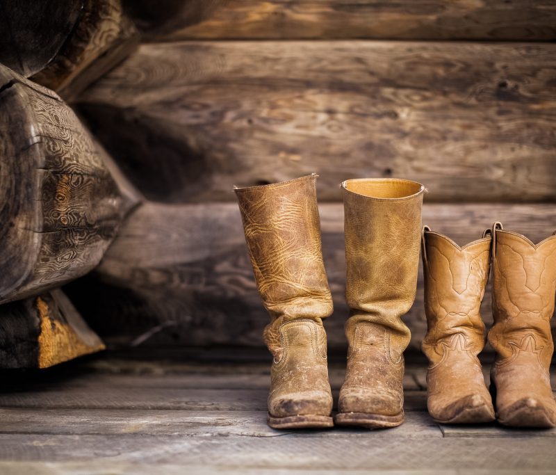 Two pairs, one big and one smaller, of rugged worn in leather cowboy boots on a barnwood floor.
