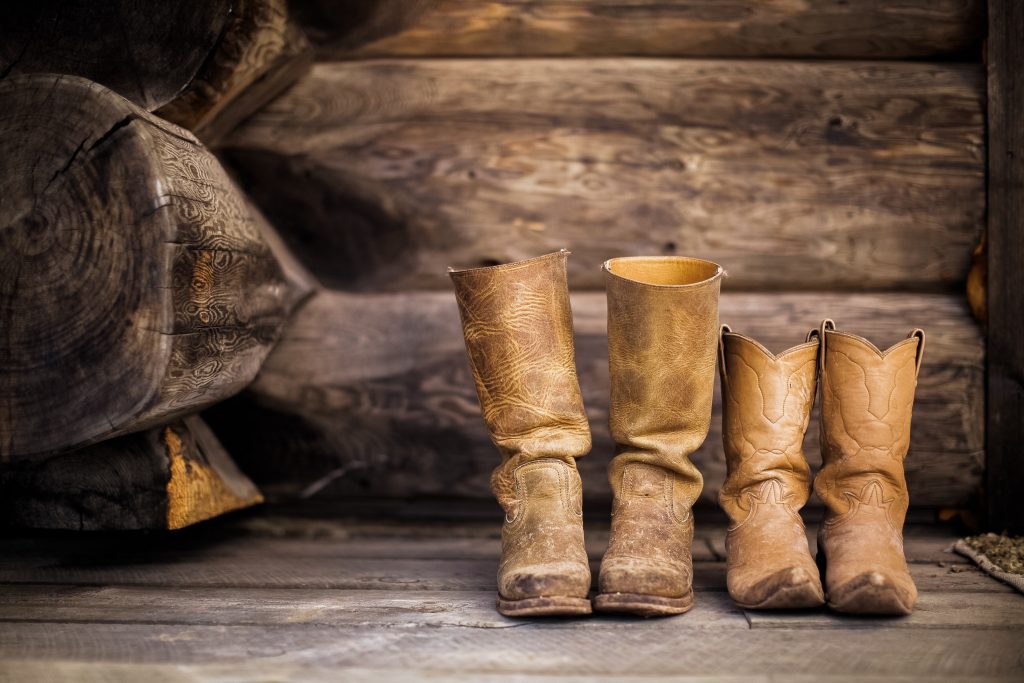 Two pairs, one big and one smaller, of rugged worn in leather cowboy boots on a barnwood floor.