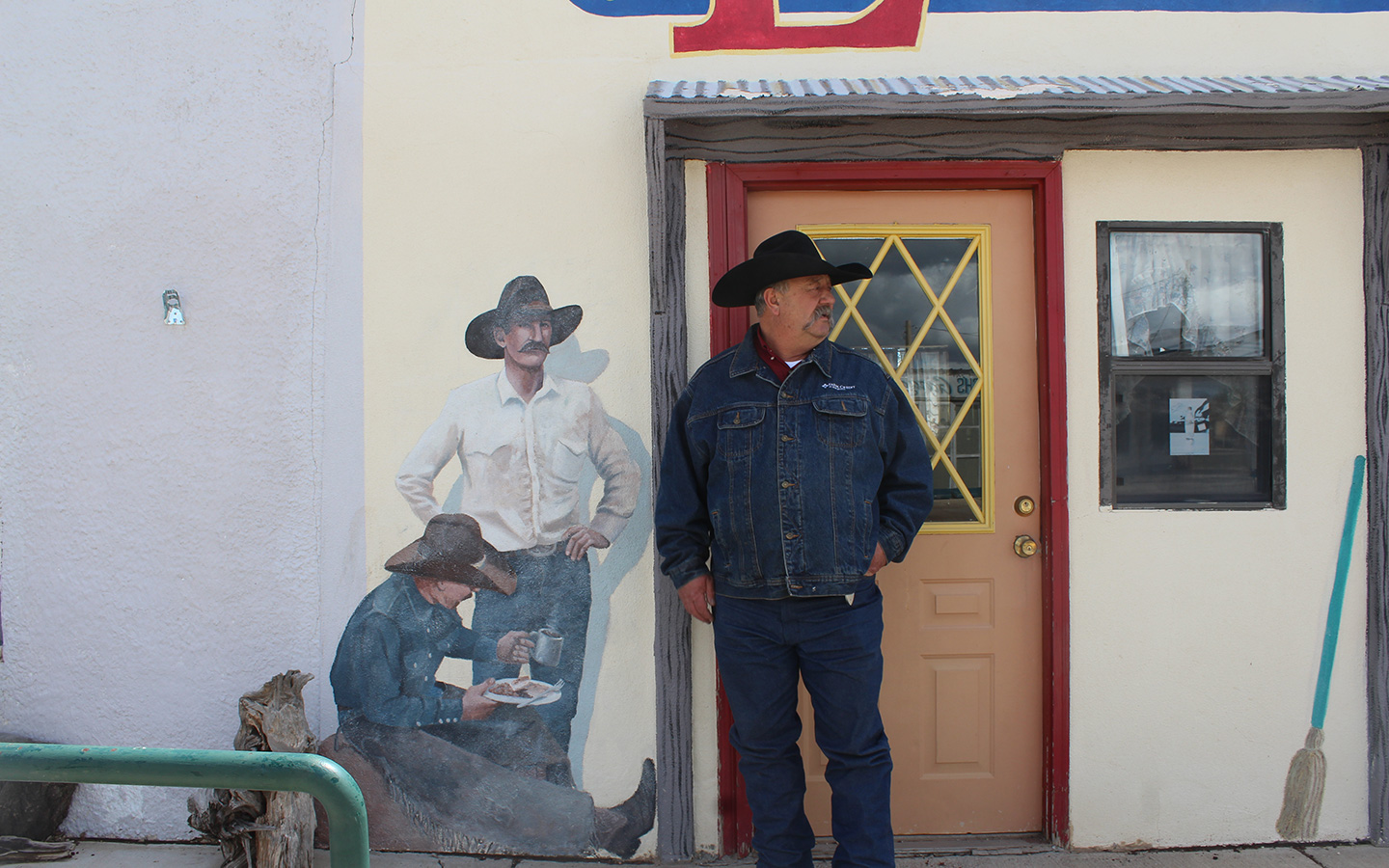 Jack Chatfield stands in front of a wall with a door behind him and mural of two men to one side.