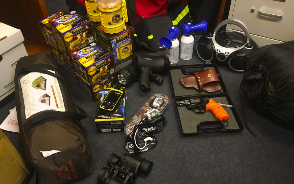 A tent, special fox lights, headlamps, a flare gun, boombox, bullhorn, and air horns, plus binoculars and a GPS device, arrayed on the carpet in an office. These items compose Lava Lake Land and Livestock's Band Kit.