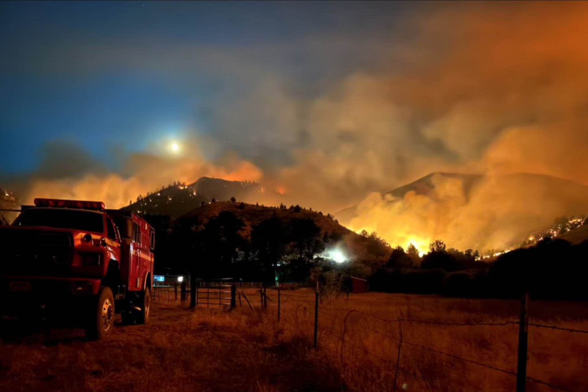 Fire truck in a field at dusk with a wildfire burning brightly in the hills just beyond.